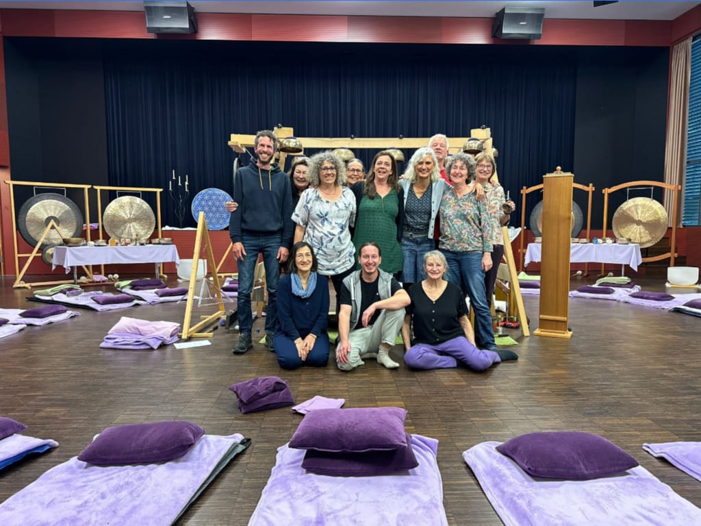 A group of people posing together in a room with musical instruments like gongs behind them. The floor has purple cushions and blankets arranged in rows. The atmosphere suggests a calm, workshop-like setting.
